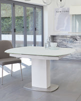 Modern dining area featuring a white oval table with a glass top on a singular pedestal base with a metal footing, paired with a taupe upholstered chair with metal legs. The setting has a minimalist aesthetic, complemented by natural light and a clean, spacious environment.