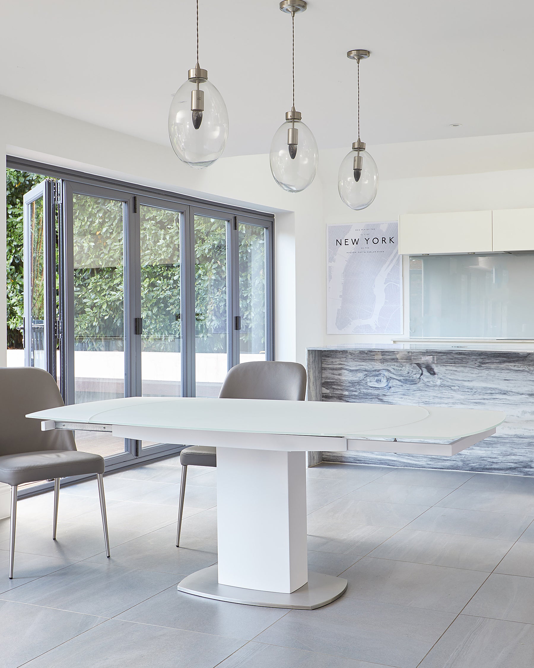 Modern minimalist dining set featuring a white oval extendable table with a pedestal base, paired with two grey upholstered chairs with sleek metal legs, set in a bright room with a contemporary aesthetic.