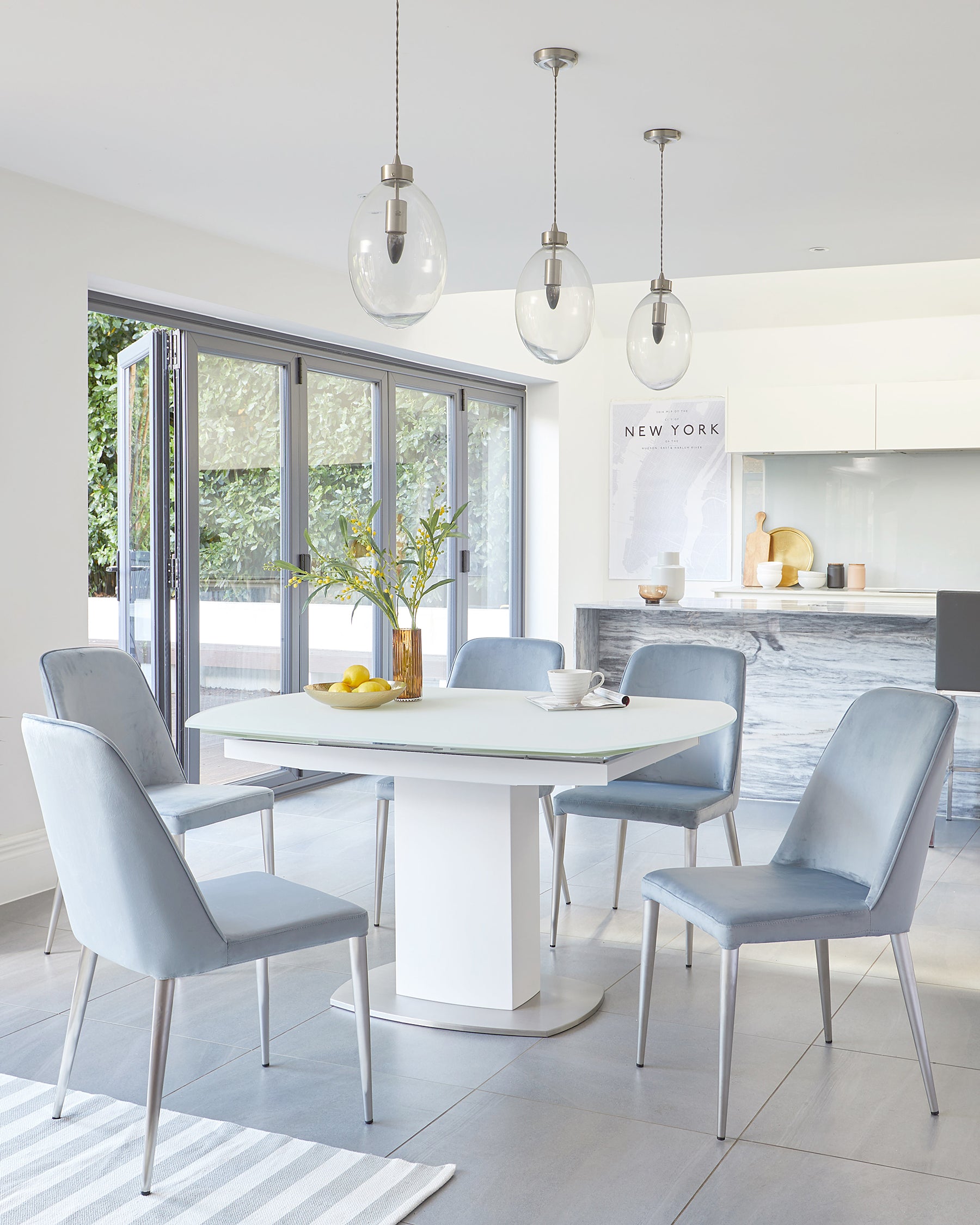 Contemporary dining room with a rectangular white dining table with sleek metal base, accompanied by four pale blue upholstered dining chairs with metal legs. The area features a striped area rug underneath the setup.