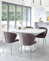 Contemporary dining room featuring a modern, white oval table with a pedestal base, complemented by four elegant, taupe upholstered chairs with sleek silver metal legs. The minimalist yet sophisticated setting is enhanced by a stylish wooden sideboard with a grey textured finish.