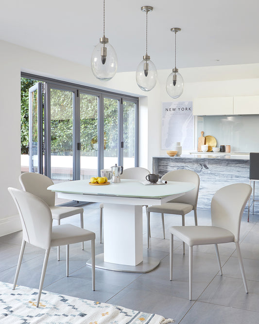 Modern dining room furniture set including a white oval dining table with a pedestal base and four beige upholstered chairs with metal legs arranged on a textured area rug.