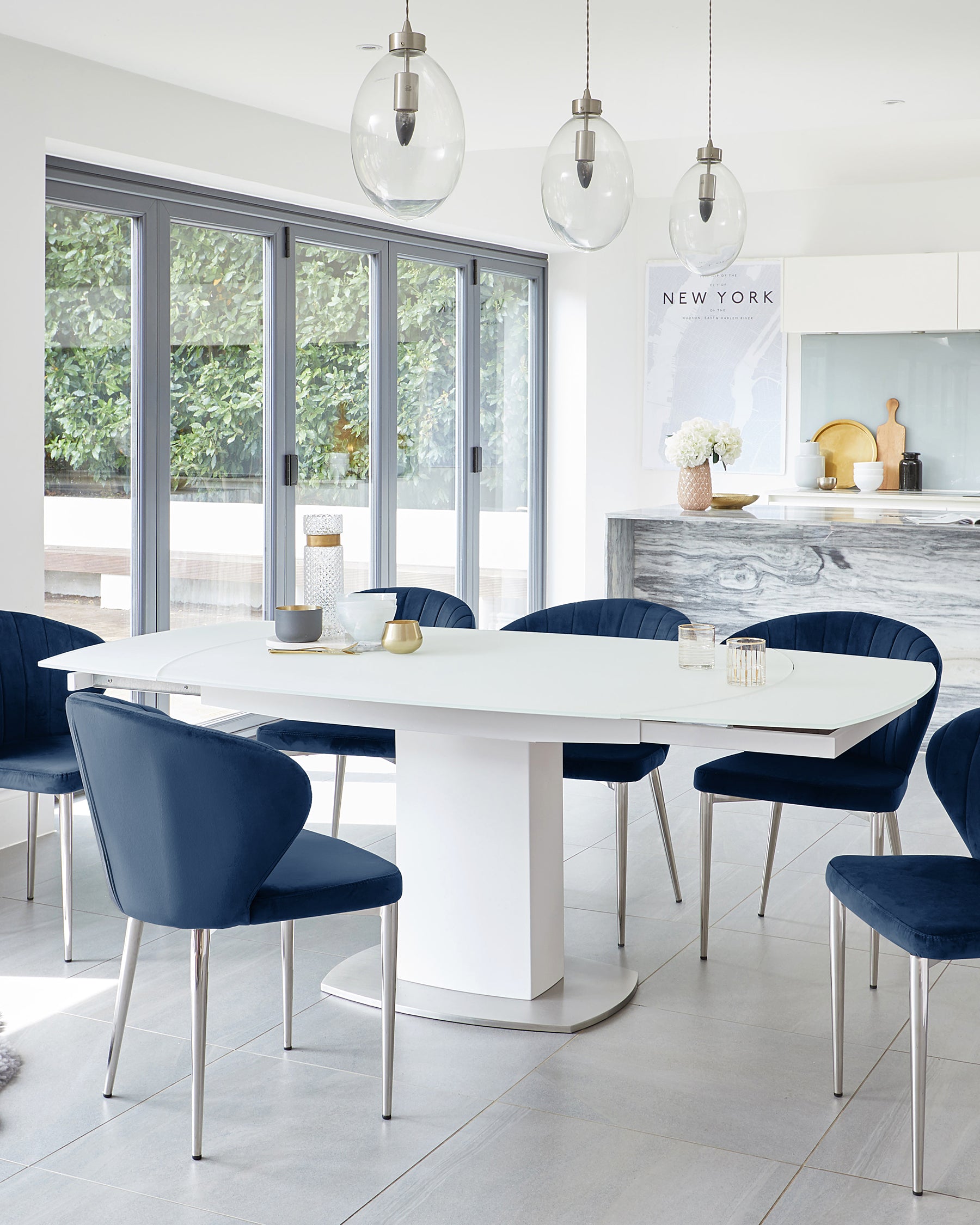 White modern oval dining table with sleek, cylindrical pedestal base, surrounded by six navy blue upholstered chairs with slim metal legs.