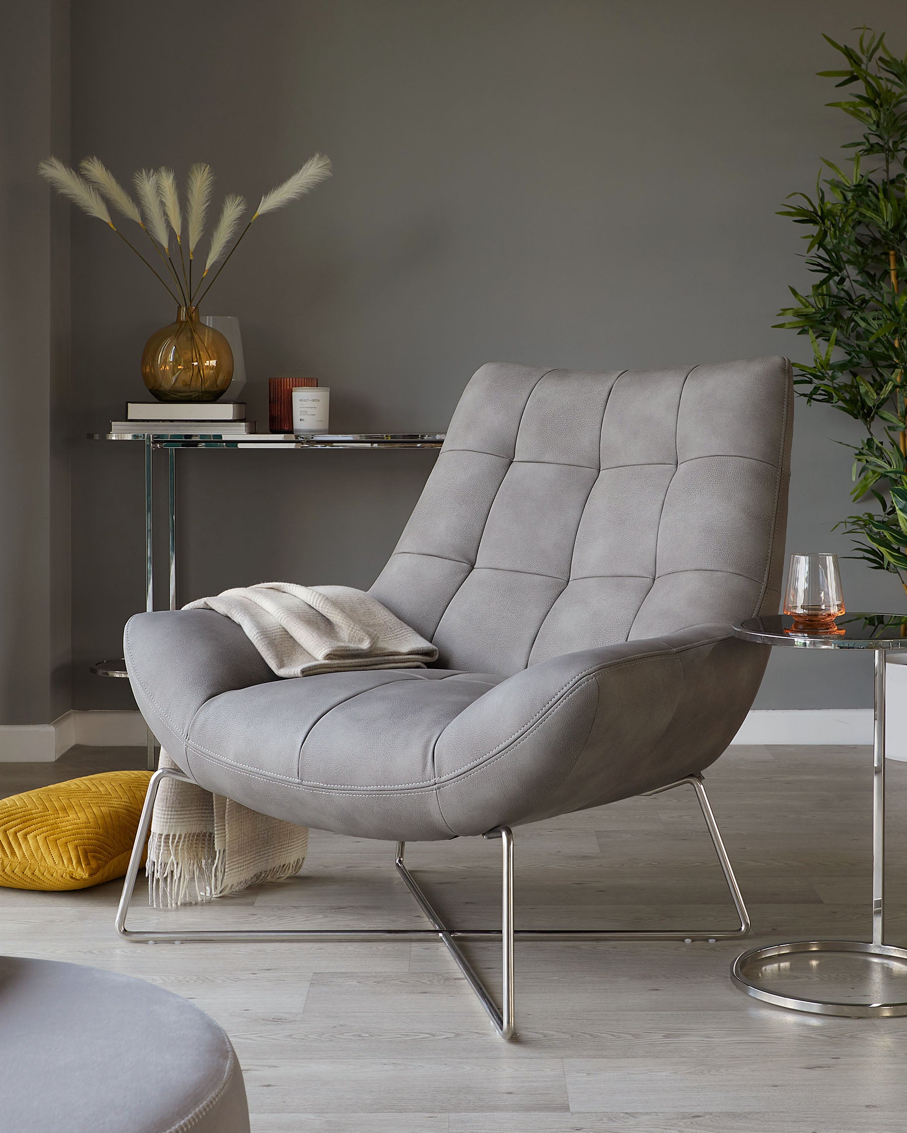 Modern grey fabric upholstered lounge chair with tufted detail and sleek chrome legs, paired with a white throw blanket and set in a contemporary living space with minimalist décor.