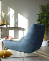 A modern blue fabric armchair with a sleek, low-back design and metal legs, accompanied by a silver minimalist side table hosting various decorative items, in a room bathed in natural sunlight adjacent to a potted plant.