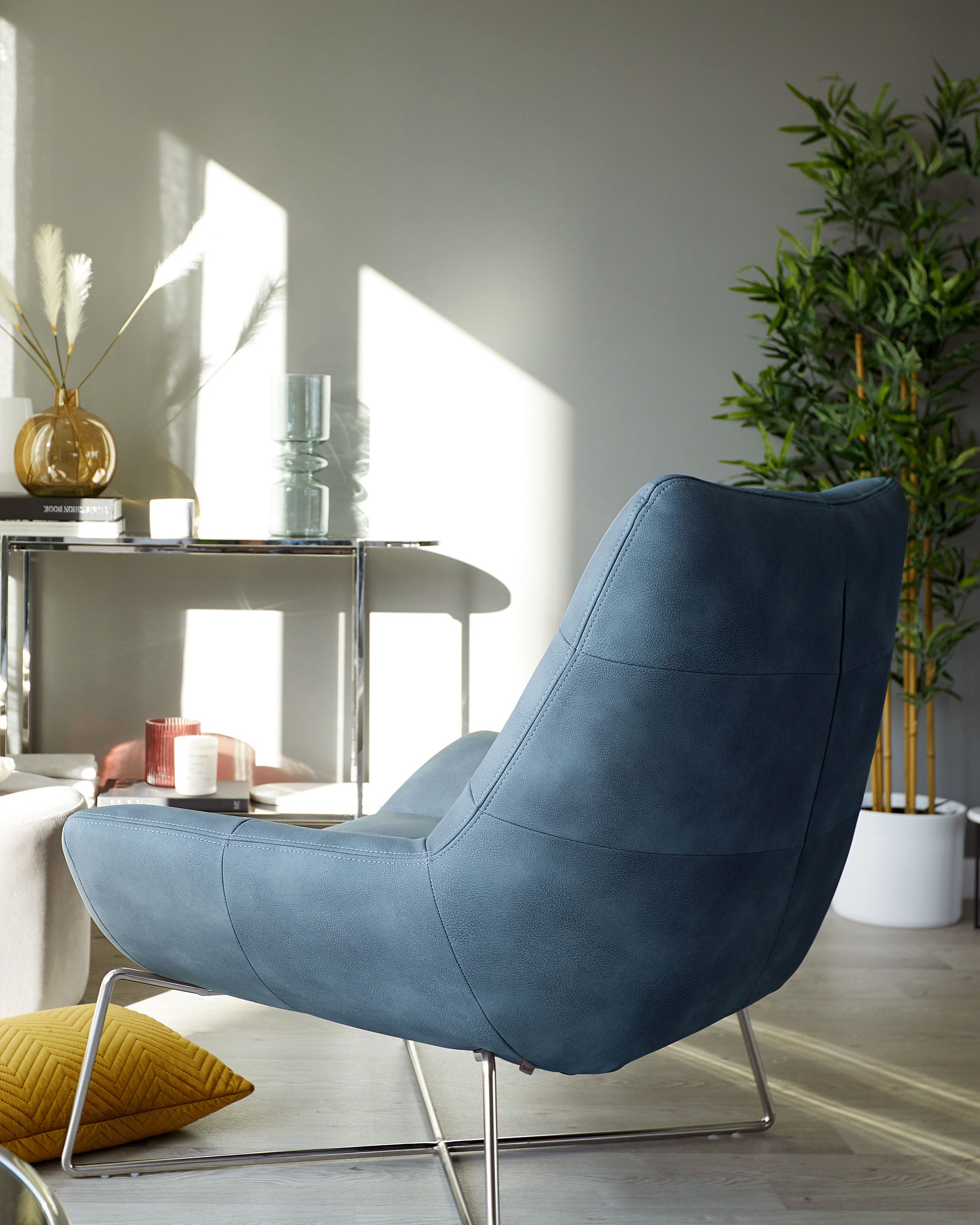 A modern blue fabric armchair with a sleek, low-back design and metal legs, accompanied by a silver minimalist side table hosting various decorative items, in a room bathed in natural sunlight adjacent to a potted plant.