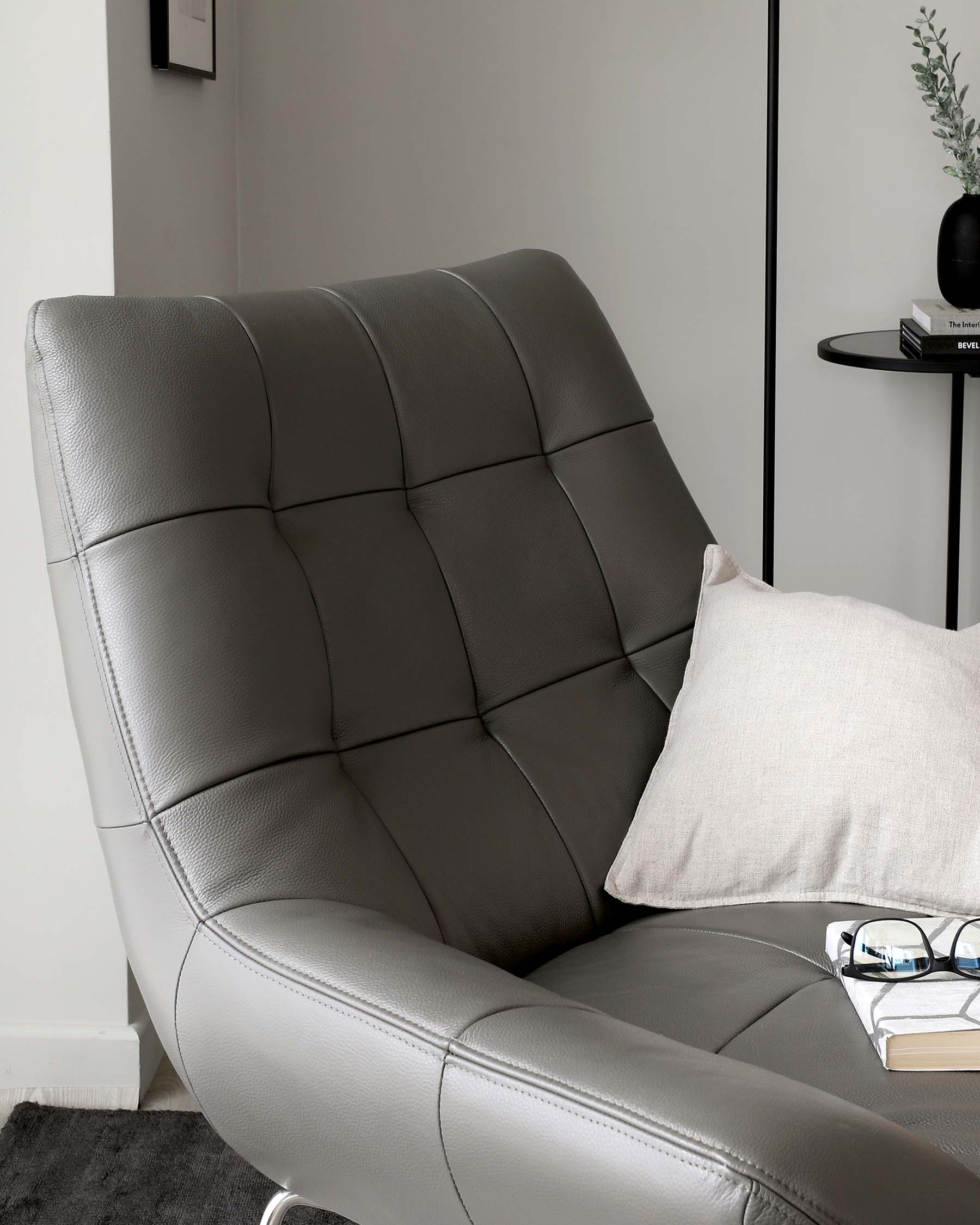 A modern grey leather recliner chair with a tufted backrest and a sleek side profile. Next to it, a minimalist round black side table holds a potted plant and a stack of books, enhancing the contemporary look of the space.