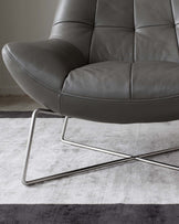 Modern grey leather lounge chair with a tufted backrest and seat cushions, featuring a sleek metal frame with a cantilever base. The chair is positioned on a two-tone grey and white area rug with a striped design.