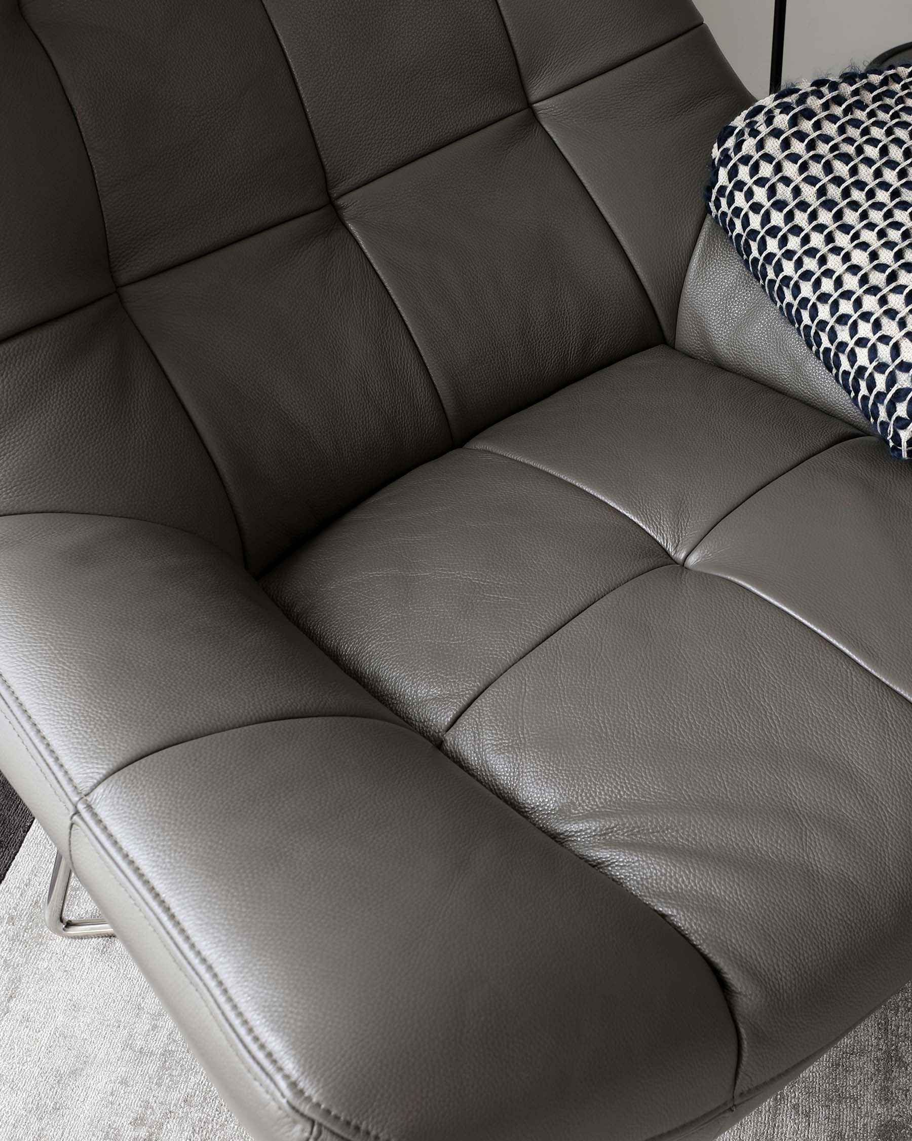 Modern charcoal grey leather recliner with tufted backrest pattern and a plush, cosy seat cushion, complemented by a geometric-patterned throw pillow on the side. The recliner has a sleek, contemporary design with a metal base, positioned on a grey textured rug.