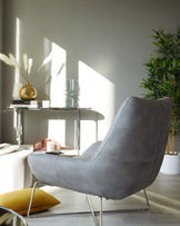 Modern grey upholstered lounge chair with a unique curving shape and sleek chrome legs, paired with a minimalist glass side table with metallic accents.