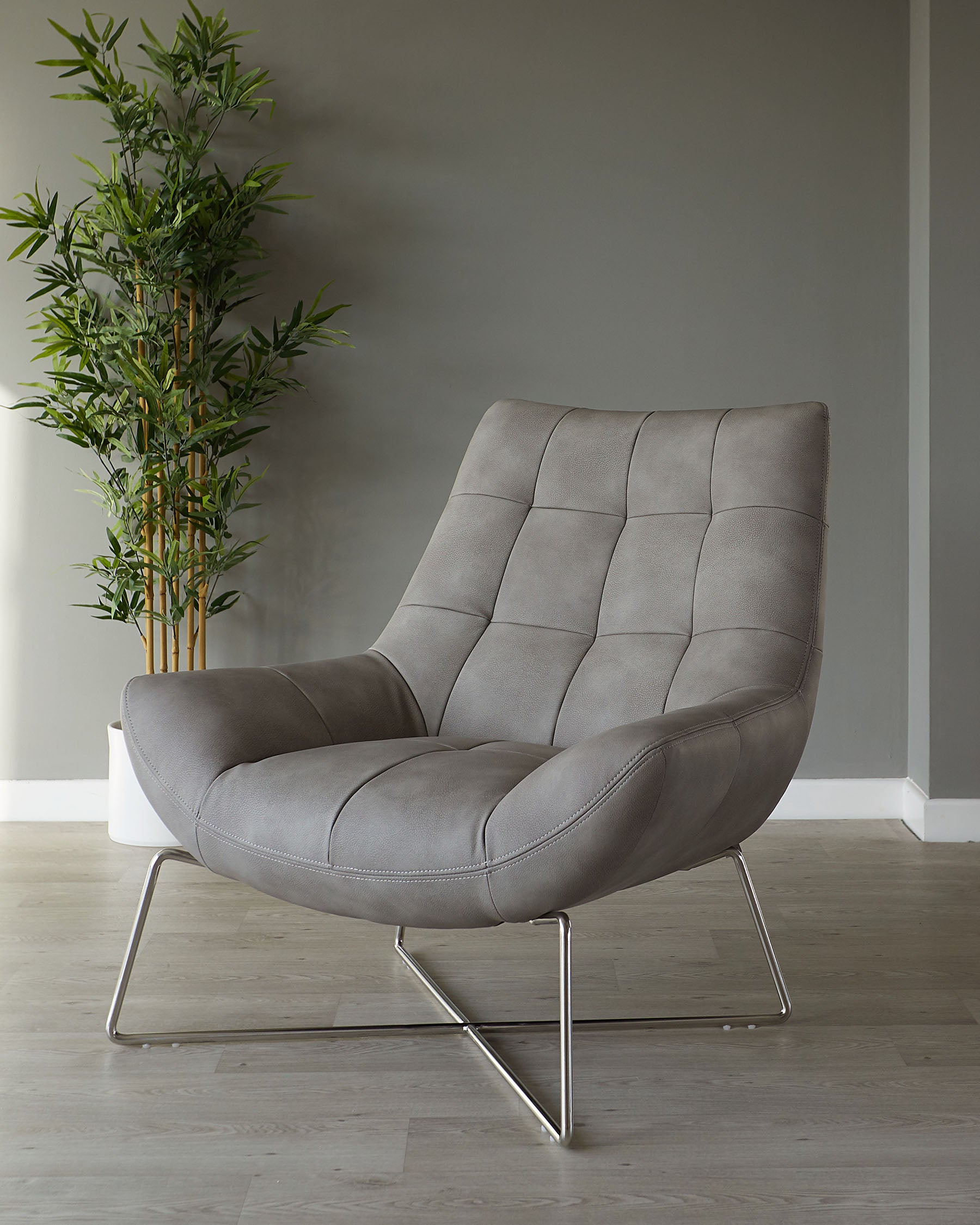 Modern grey upholstered accent chair with tufted backrest and cushioned seat, supported by sleek silver metal legs in a geometric design, staged on a light wood floor with a bamboo plant in the background.