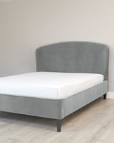 Grey upholstered platform bed with a curved headboard and simple dark wooden legs, showcased in a room with light wooden flooring and a neutral wall. A white mattress is placed on top of the bed frame.