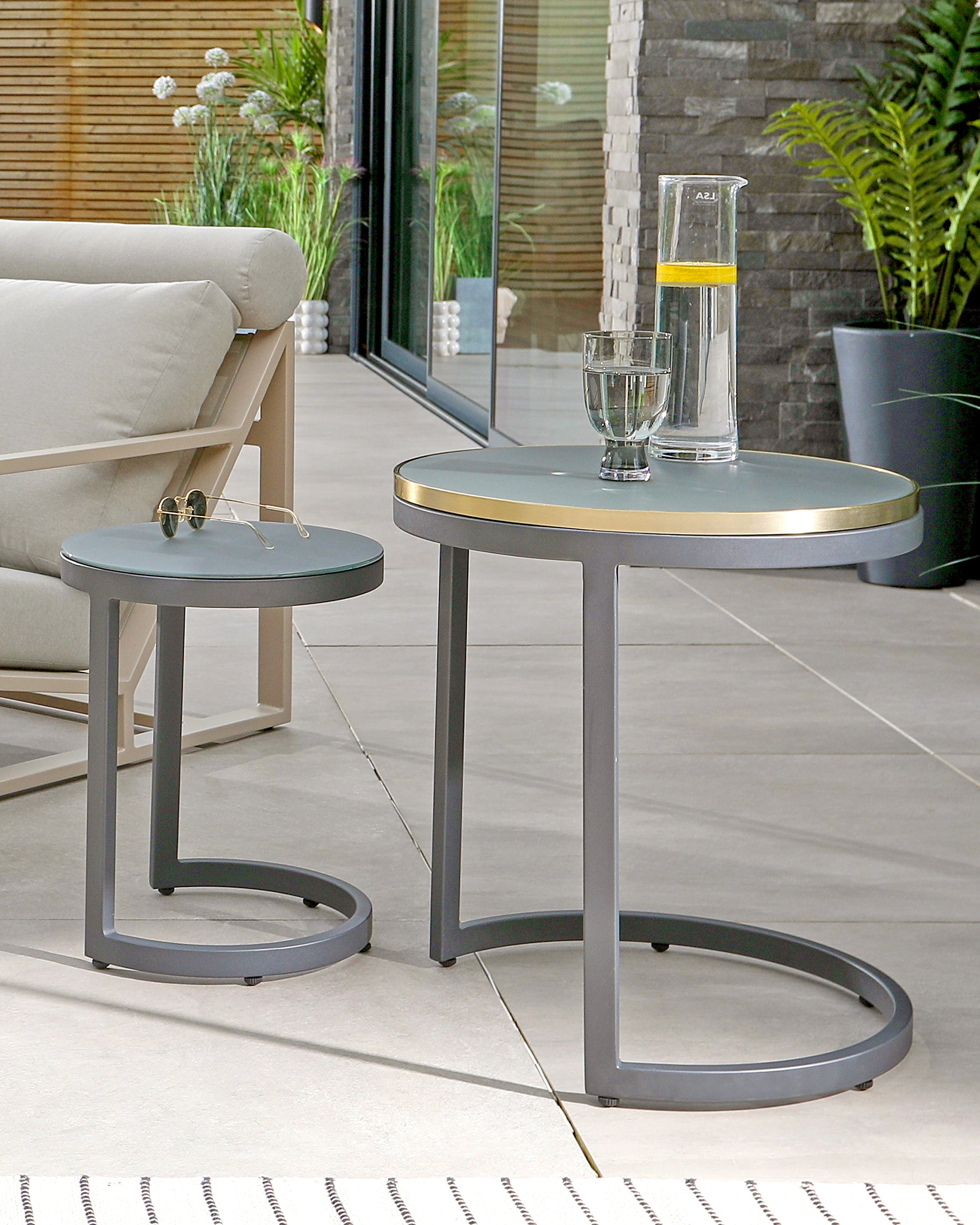 A set of two contemporary round side tables with slender metal frames and smooth, matte-finished tabletops. The larger table features a brass edge banding accent, while both stand on sleek circular bases that ensure stability on the tiled patio floor. A water carafe and glass rest atop the larger table, with a pair of sunglasses on the smaller one, hinting at casual outdoor leisure.