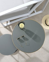 Modern minimalist furniture set featuring a white wooden bench with light grey cushions and a sleek round side table with a glass top and gold trim. A smaller glass accent table echoes the design beneath the main table, contributing to a sophisticated layered look.