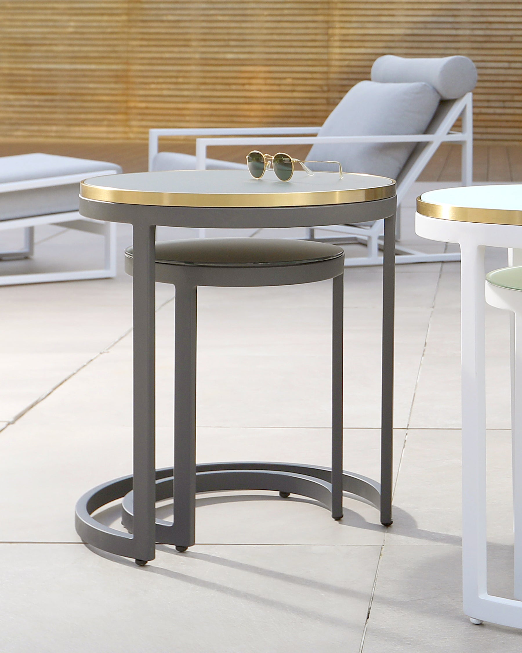 Contemporary outdoor furniture arrangement including a round side table with a metallic gold top and grey frame accompanied by a coordinating stool with a black seat and grey legs. In the background, there is a glimpse of a white-framed lounge chair with light grey cushioning.