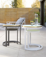 Modern outdoor furniture with a white lounge chair in background, and three nesting side tables with round tops in black, white, and metallic finishes in the foreground. One table holds a glass and a clear water carafe.