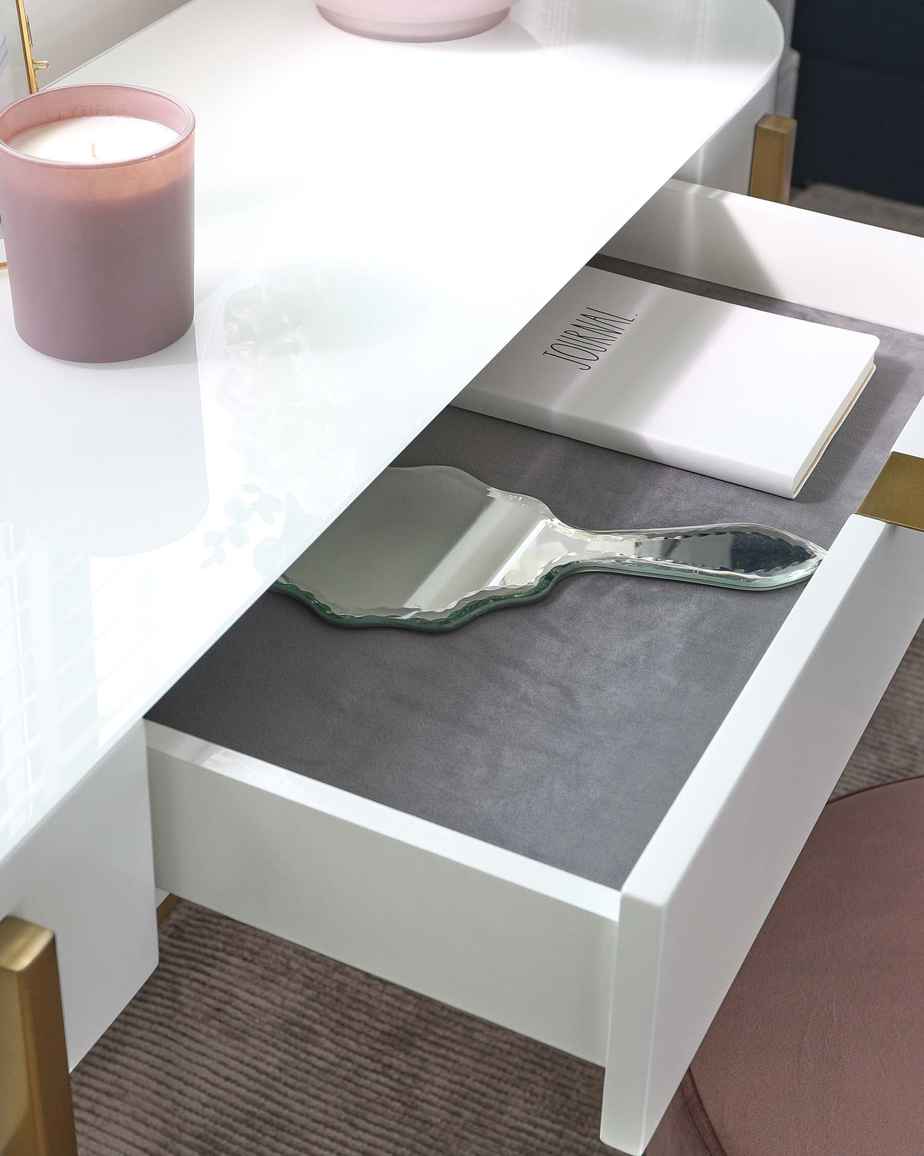 Modern white glossy coffee table with an open drawer featuring a built-in grey felt liner, flanked by brass-coloured table legs, accessorized with a large scented candle, a hardcover journal, and a decorative glass tray.