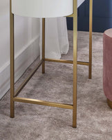 Modern minimalist furniture featuring a round white top side table with a sleek golden metal frame and a cylindrical blush pink upholstered ottoman with a plush velvet finish.