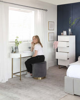 A modern bedroom with a sleek white floating desk paired with a circular grey velvet stool featuring a gold base. The room includes a tall white dresser with clean lines and simple handles, and a light grey upholstered bed with a minimalistic headboard. The furniture is complemented by a pale grey area rug and accented with understated wall art and plants.