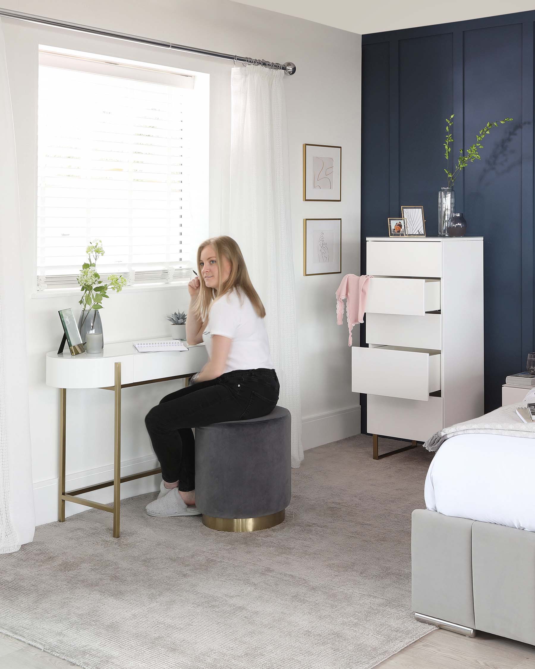 A modern bedroom with a white semi-circular desk with a gold metal base, paired with a round, grey velvet ottoman featuring a gold band at the bottom. Beside the window, there's a tall white chest with asymmetric shelves, displaying books and a vase of greenery. The room has a minimalist grey upholstered bed partially visible in the foreground.