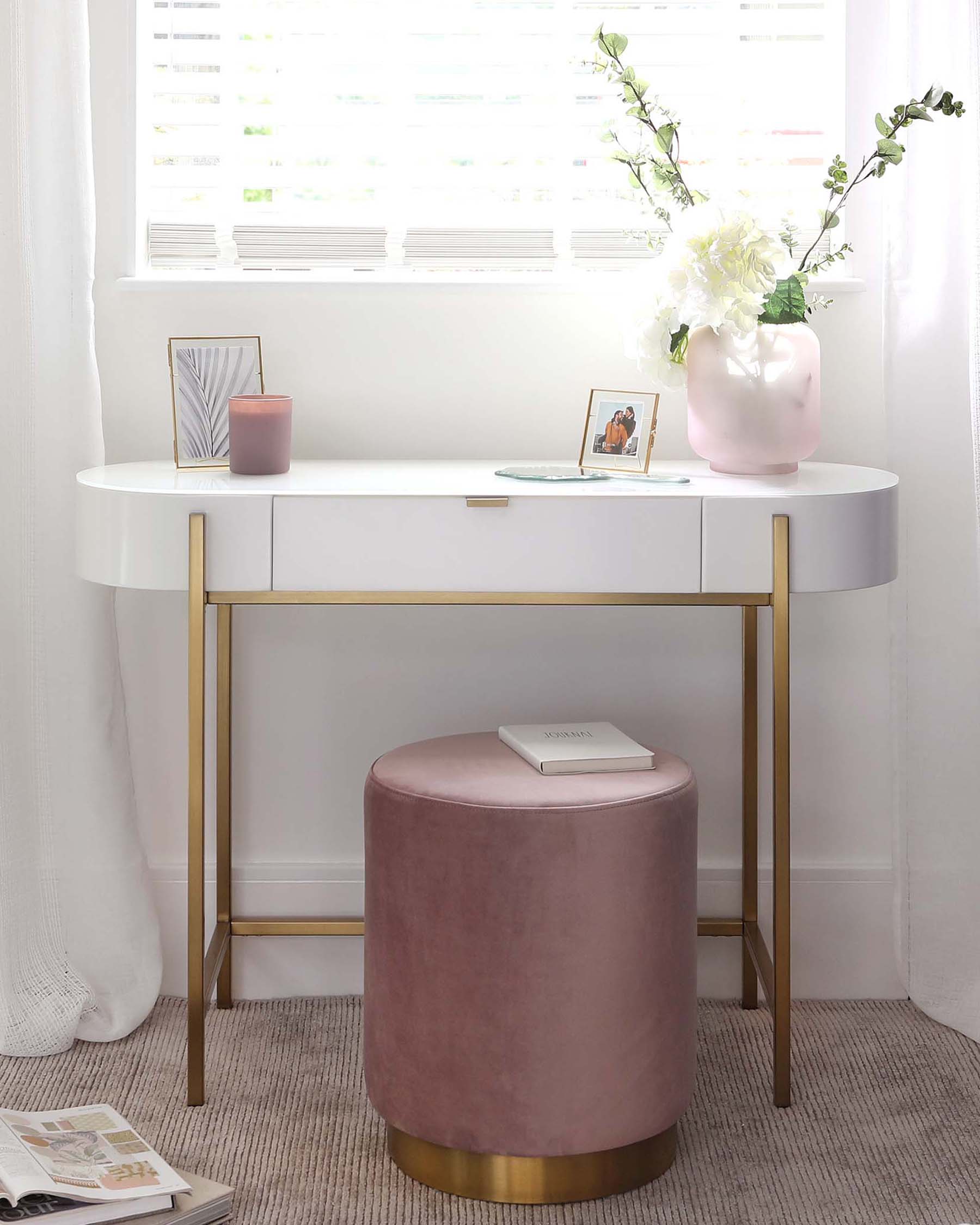 Elegant modern furniture set including a white oval desk with a gold metal base and a pink velvet round ottoman with a gold metal band at the base. The desk displays minimalistic decor, and the ottoman has a book on top, suggesting a chic and functional design suitable for a contemporary interior.