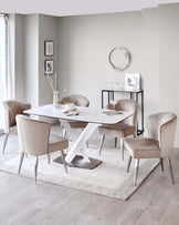 Modern minimalist dining room featuring a white rectangular table with a unique X-shaped metallic base, surrounded by four plush, velvet upholstered chairs in a soft beige tone with sleek metallic legs. A small, squared side table with a glass top and thin metal frame stands near a wall, adorned with frames and a vase. The room is completed with a circular mirror on the wall and a neutral-toned rug under the dining set.