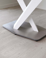 Modern cross-legged table base with a white finish and brushed metal rectangular platform, set on a light wooden floor.