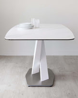 Modern minimalist dining table with a white marble-effect top and a unique geometric metal base in a white and silver finish, set against a light hardwood floor and a neutral wall, with a simple set of white plates and clear glasses on top.