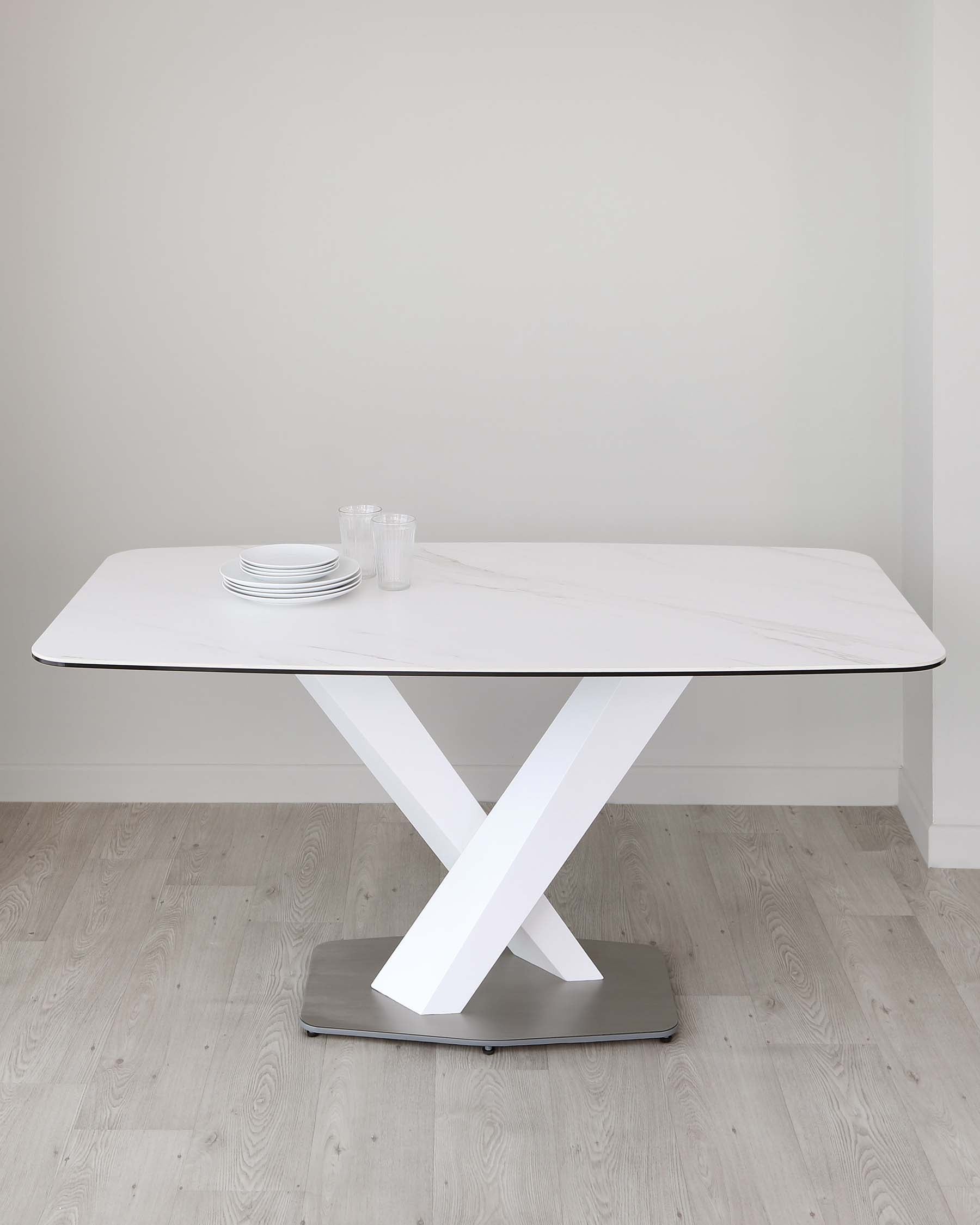 Modern rectangular dining table with a white marble top and a unique white X-shaped base, set on a flat grey platform. The table is accessorized with a stack of white plates and clear drinking glasses. The setting is minimalist with a neutral background and light wooden flooring.