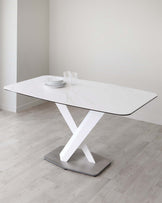 Modern rectangular dining table with a white marble top and a striking geometric white and metallic base, set against a clean, minimalist background. On the table, there is a simple setting of white plates and clear drinking glasses.