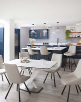 Modern dining room with a white oval table featuring a unique x-shaped base and complemented by six light grey upholstered chairs with sleek black legs. In the background, a blue kitchen island with bar stools showcases a matching contemporary design.