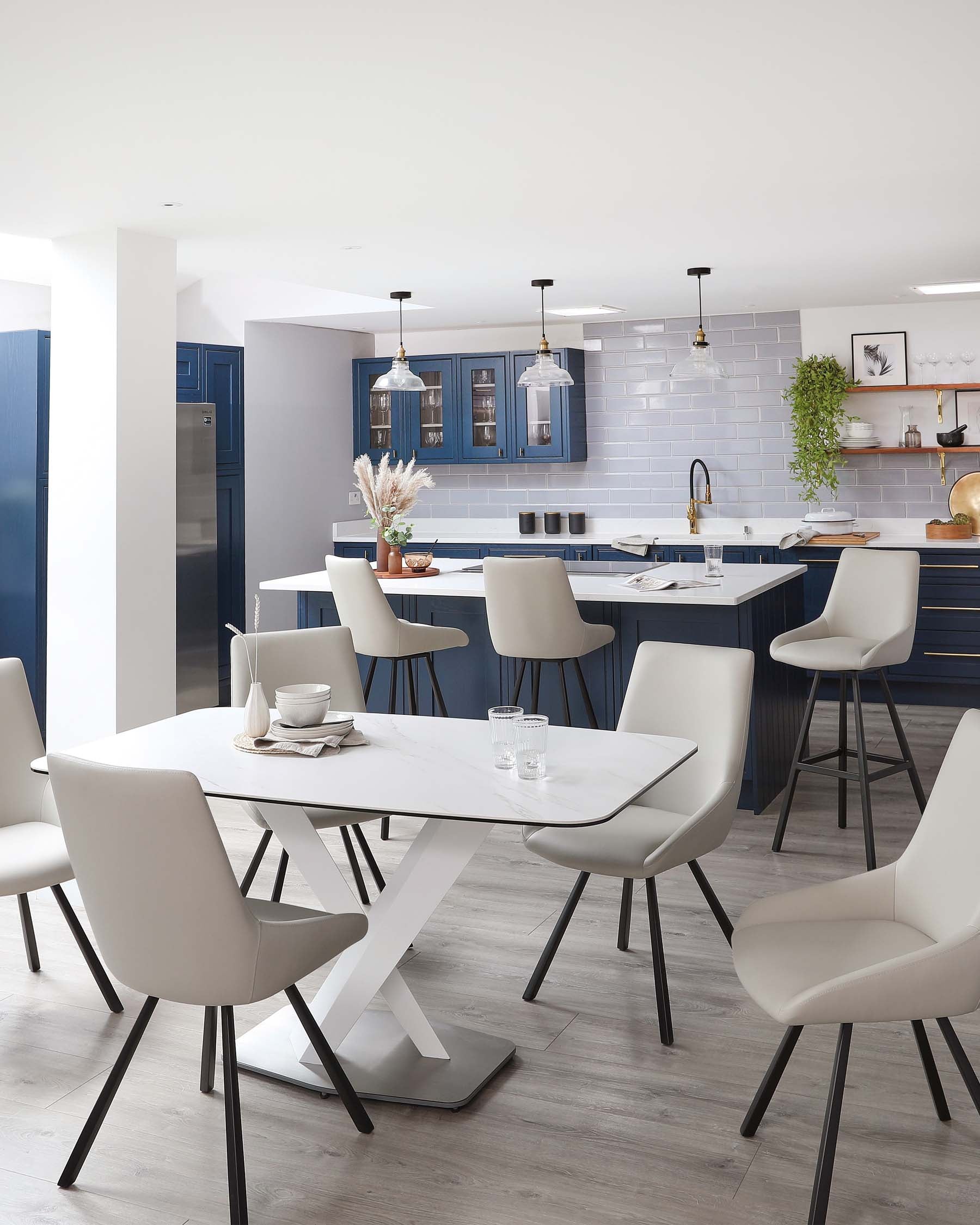 Modern dining room furniture including a white oval dining table with a unique X-shaped base and six contemporary light grey upholstered chairs with slim black metal legs. A kitchen island with a white countertop and bar stools with dark blue upholstery and black frames is visible in the background. The pendant lighting above the island and table contributes to the overall sleek and minimalist aesthetic of the space.