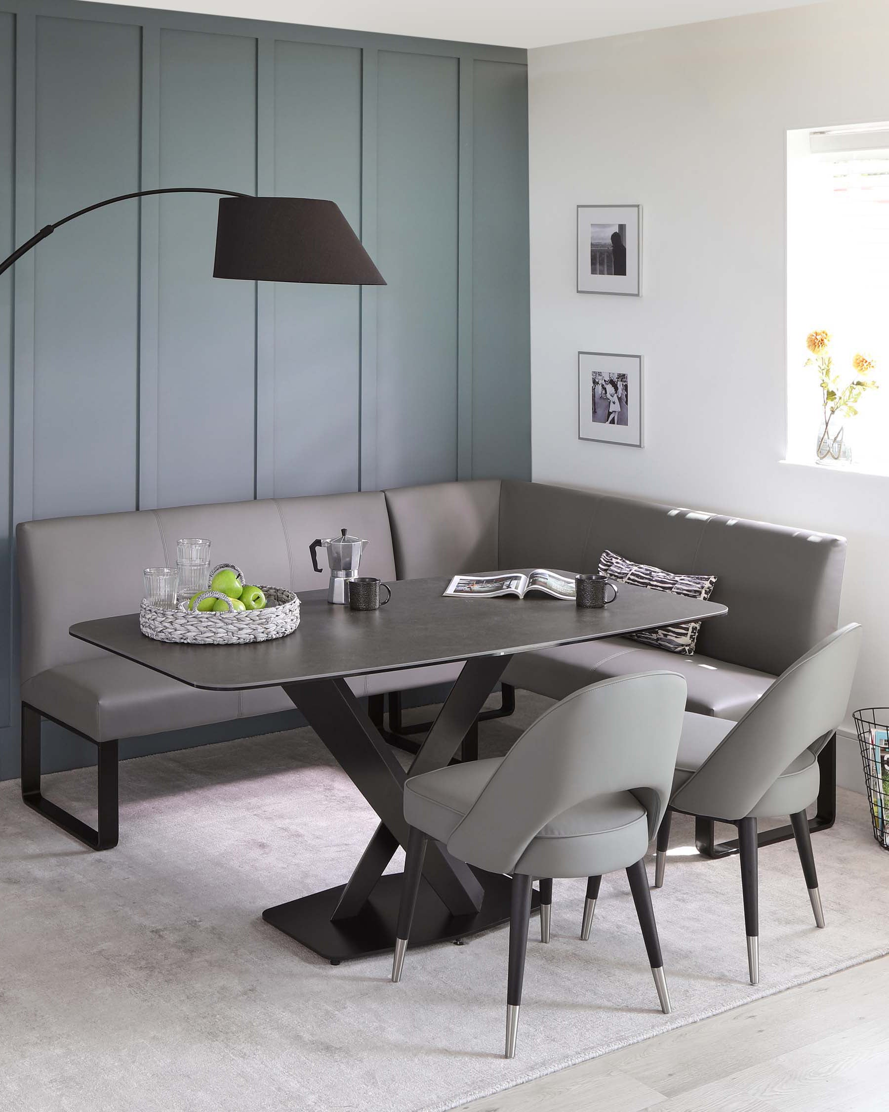 Modern dining set featuring a rectangular table with a dark stone finish and unique geometric black base. Accompanied by elegant grey upholstered chairs with black legs and a matching grey corner bench under a sculptural black floor lamp.