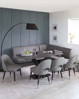 Modern dining room furniture set featuring a sleek, oval-shaped black dining table with clean lines, a built-in grey corner bench upholstered in leather-like material, and five matching grey dining chairs with curved backs and metal legs. A complementary arched floor lamp with a large black shade extends over the table, enhancing the contemporary aesthetic.