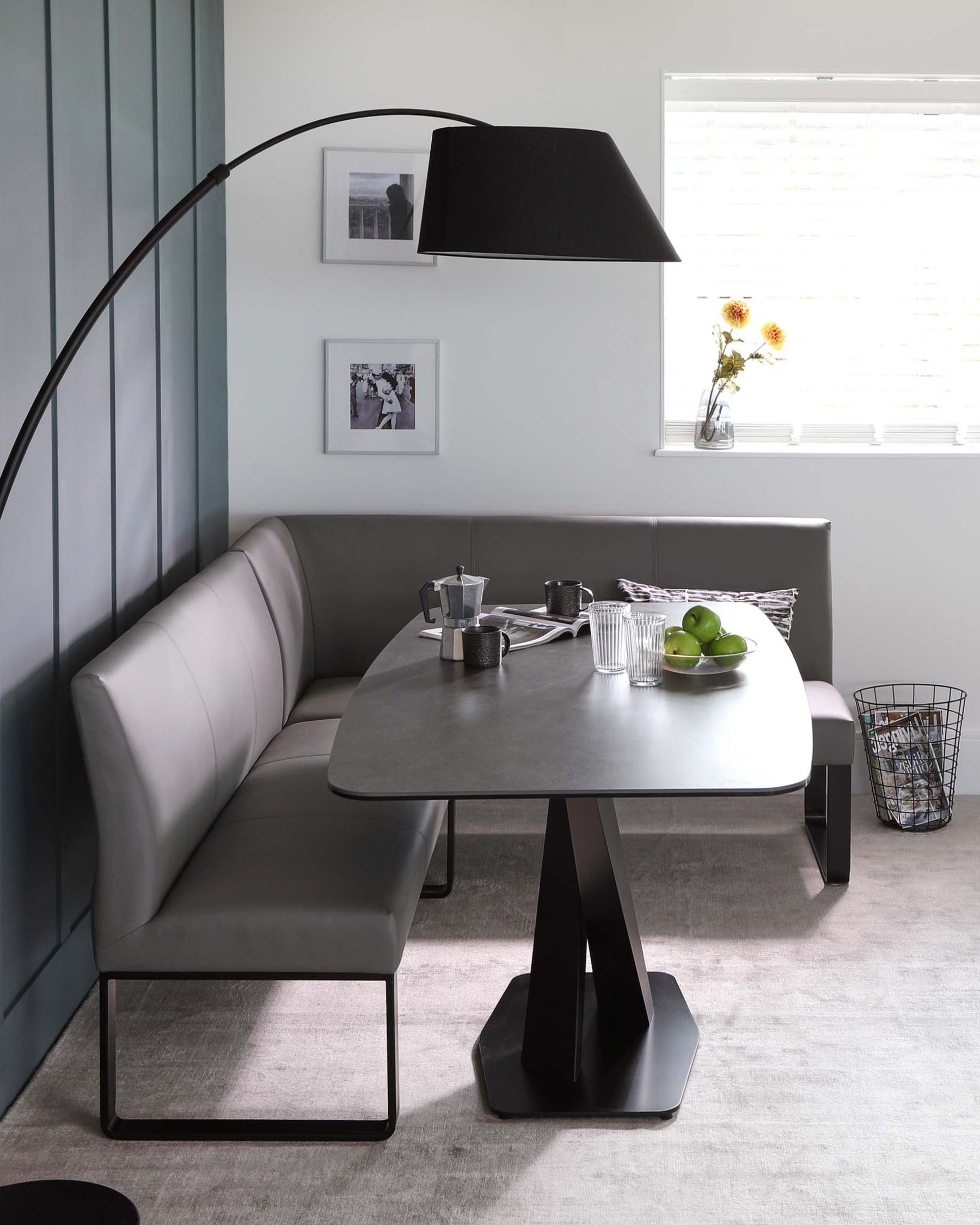 Modern dining space featuring a sleek, contemporary rectangular table with an elliptical top and geometric triangular pedestal base in a matte finish. Paired with a corner bench upholstered in a light grey leather-like material, the ensemble is completed by a striking oversized arched floor lamp with a large black shade, illuminating the area. A casual wire trash basket adds to the room's minimalist aesthetic.