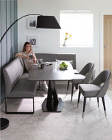 Modern dining set featuring an oval table with a matte black pedestal base and a grey tabletop, accompanied by sleek, grey fabric upholstered dining chairs with slim metallic legs. A curved floor lamp with an oversized black shade arches over the ensemble, adding a contemporary touch to the minimalist space.