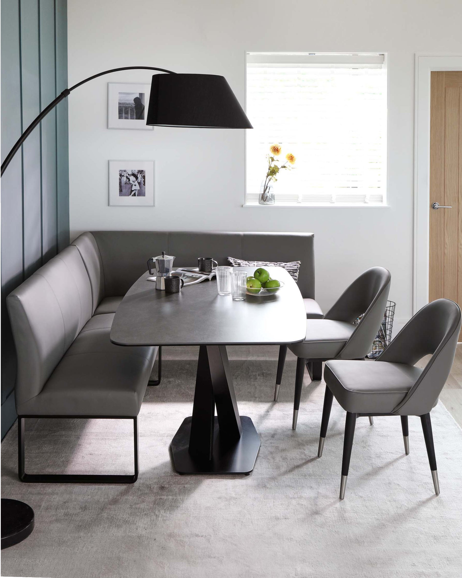 Contemporary dining room furniture featuring a grey corner bench with a high backrest, along with a sleek, dark grey table with an unusual geometric base. It is complemented by two chairs with grey upholstery and black metal legs, set on a textured light grey area rug. A modern black floor lamp arches over the ensemble.