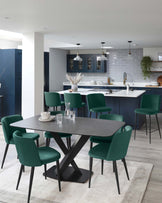 A contemporary dining set featuring a sleek, oval-shaped table with a dark top and a striking X-shaped black base, paired with six plush, green upholstered chairs with black legs. The room has a modern aesthetic with a neutral palette and wood flooring.