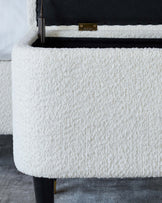 Close-up of a textured white fabric upholstered storage ottoman with a hinged lid partially open, revealing black interior lining, set on slender black legs tipped with gold accents.