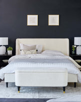 Elegant contemporary bedroom furniture set featuring an upholstered queen-size bed with a textured cream headboard and matching bench at the foot, both with black legs that include brass cap accents. A plush grey area rug underlies the bed and bench ensemble.