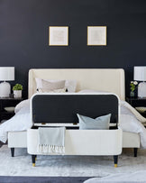 Elegant contemporary bedroom furniture featuring an upholstered king-size bed with cream textured fabric and a tufted headboard, flanked by two matching white ceramic table lamps on minimalist nightstands. At the foot of the bed is a bench with the same cream upholstery and black lined accents, raising on black legs with gold feet caps. The room is accessorized with a variety of pillows, a chevron-patterned throw blanket, and subtle artwork above the bed.