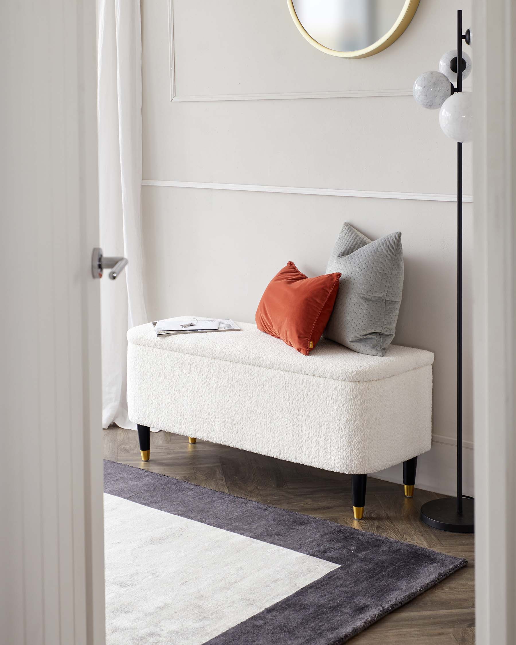 Elegant off-white upholstered bench with textured fabric, featuring black and gold tapered legs, accessorized with two square throw pillows in contrasting red and grey tones. A sleek black floor lamp with spherical white details stands beside the bench, all atop a contemporary geometric area rug with a gradient of white to dark grey. A round gold-framed mirror hangs on the wall and a casual magazine rests upon the bench, suggesting a cosy reading nook.