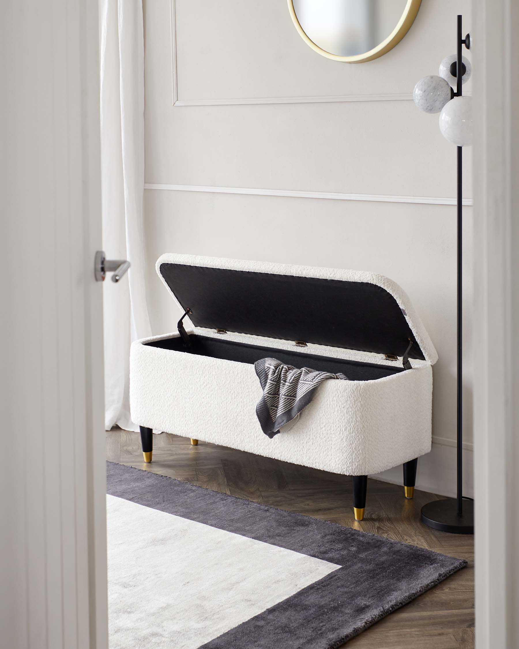 Elegant modern storage bench with a white bouclé upholstery and black interior, featuring tapered legs with gold-finished caps. A striped throw blanket is casually draped on one side, and the bench is positioned on a geometric patterned area rug in monochrome tones. Behind the bench is a minimalist floor lamp with marble accents, and a round gold-rimmed mirror hangs on the wall, enhancing the room's sophistication.