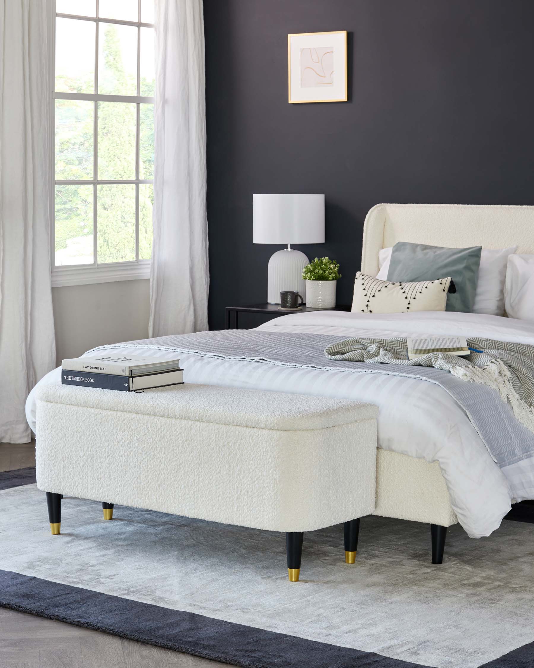 Elegant bedroom scene featuring a plush, white textured upholstered bench with sleek black legs tipped with gold caps at the foot of a neatly made bed with striped and solid pillows. A dark wood nightstand with a ribbed white ceramic lamp is to the side, against a dark grey wall with a minimalist line art frame. Flowing white curtains frame a window with a view of greenery, completing the serene and sophisticated decor.