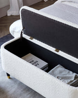 Modern dual-purpose storage bench with a plush, off-white textured upholstery exterior and black fabric interior. The bench is open to reveal ample space with a book and neatly folded throw blanket inside. It features black fabric on the underside of the lid, black lining inside, and is finished with stylish, slender brass-coloured metal legs.