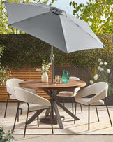 A round wooden table with a dark metal base, surrounded by four light grey upholstered chairs under a large grey patio umbrella.