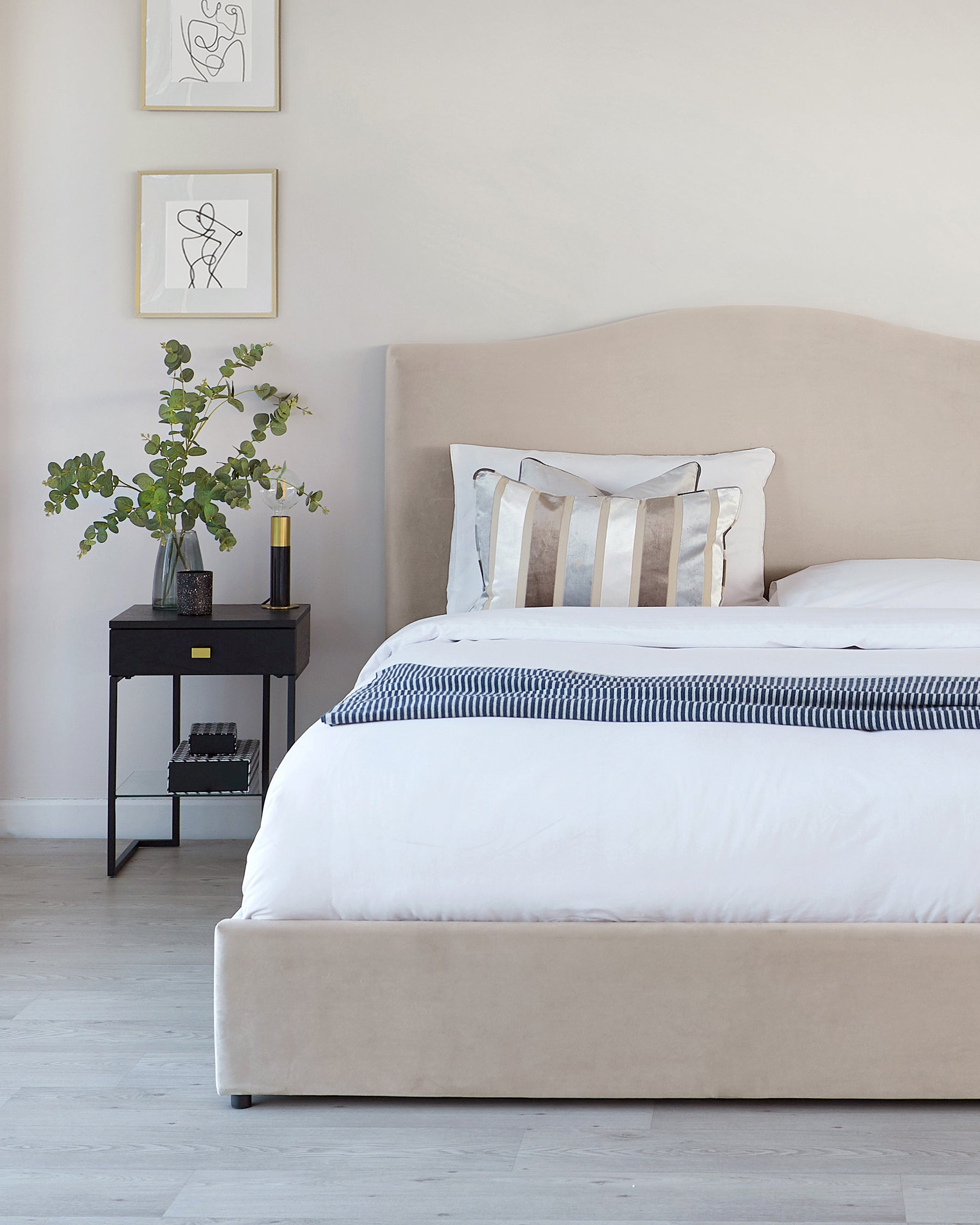 Elegant bedroom featuring a contemporary beige upholstered bed with a gently curved headboard and minimalistic black metal side table with square top, lower shelf, and slender legs. A decorative potted plant and modern black accessories adorn the side table.