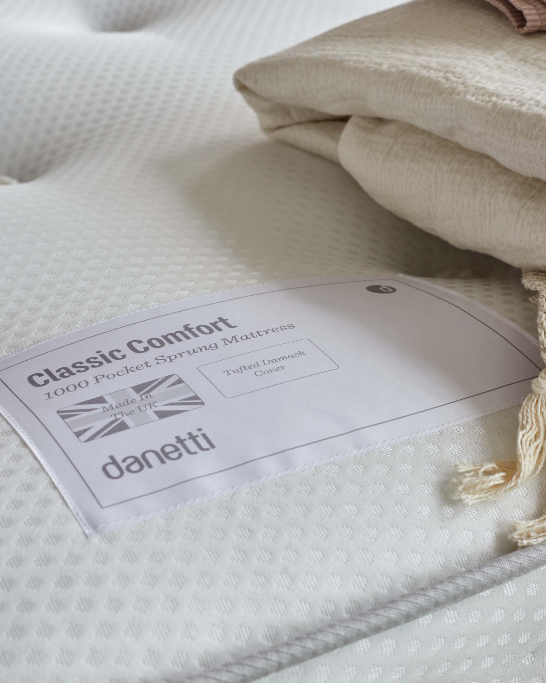 Close-up of a textured white pocket sprung mattress with an attached label reading "Classic Comfort 1000 Pocket Sprung Mattress," accompanied by a folded natural-toned blanket on top.