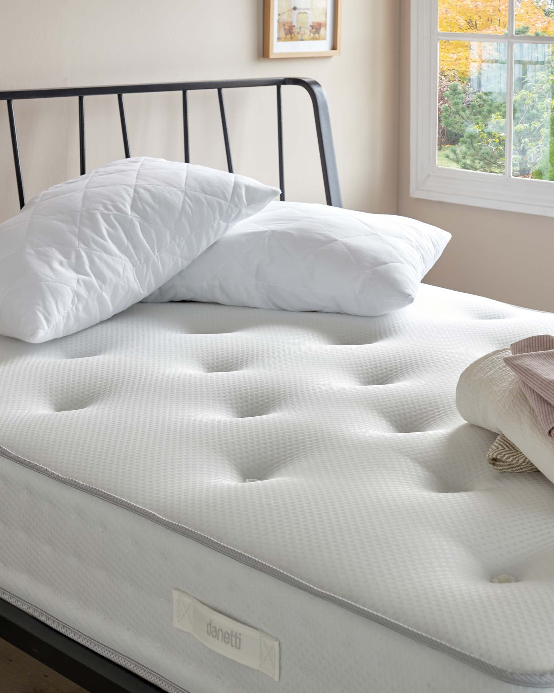 A modern minimalist black metal bed frame with a white quilted mattress. The bed is adorned with a pair of white pillows and positioned near a window with a view of autumn foliage.