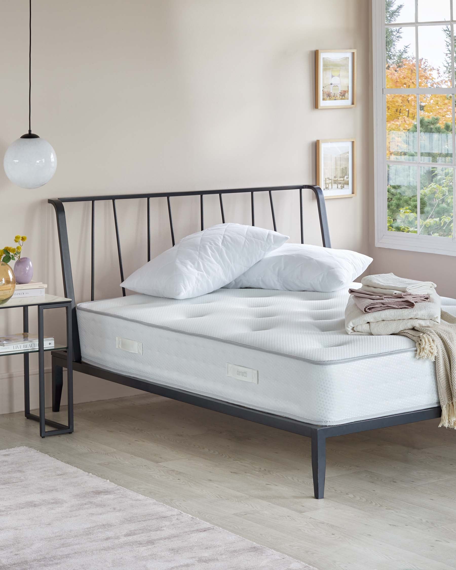 A contemporary bedroom scene featuring a black metal bed frame with a simplistic headboard design, a white mattress, two fluffy pillows without cases, and a neutral-toned throw blanket casually folded at the bed's foot. Adjacent is a minimalist black metal side table with a square tabletop and a single shelf underneath.