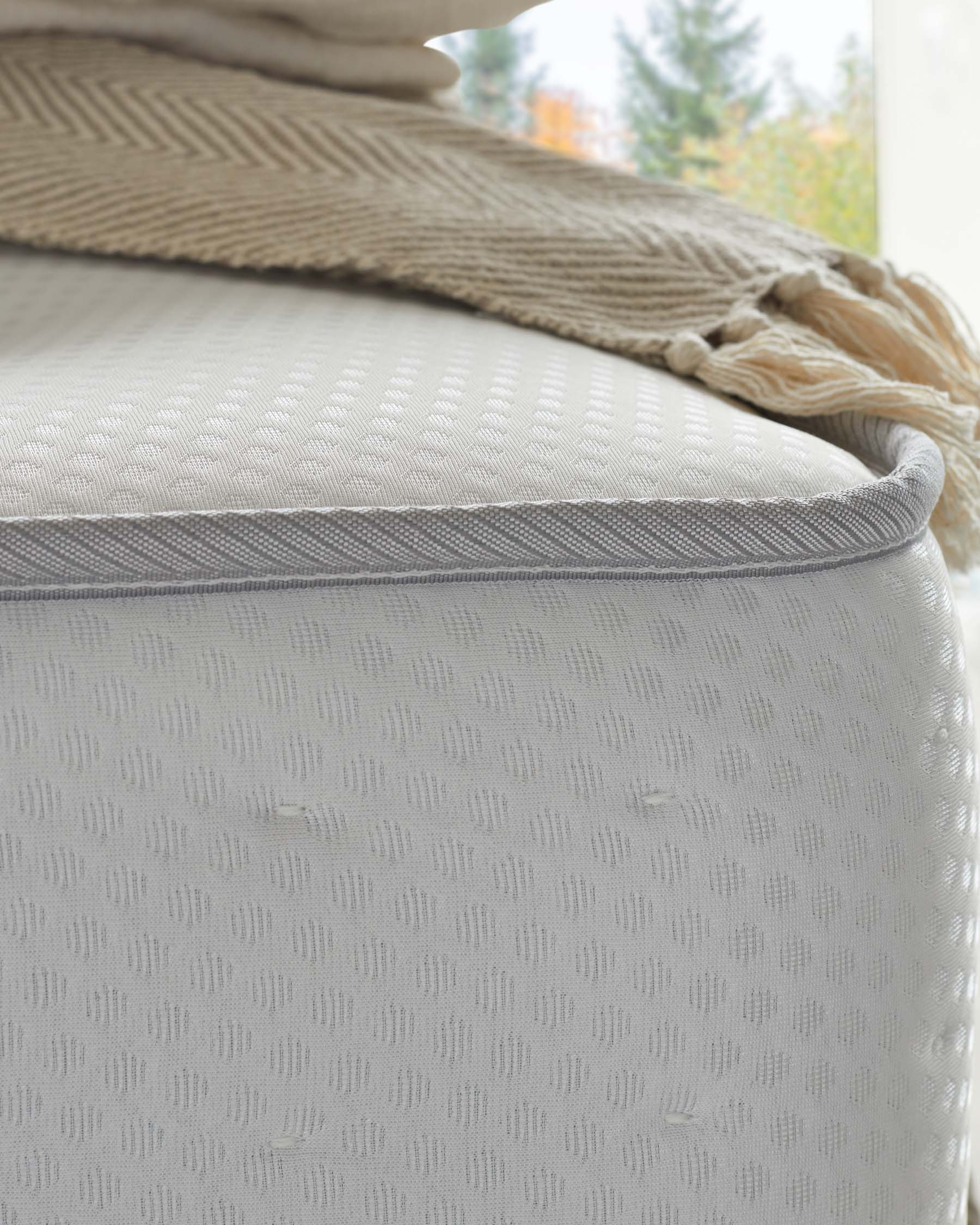 Close-up of a part of a soft white fabric upholstered mattress with a herringbone knit beige throw casually draped across the top, showcasing a cosy and elegant bedding arrangement near a window with natural light.